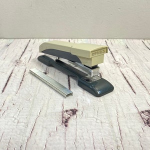 Può includere: Una graffatrice grigia e beige con un estrattore di punti metallici argentato. La graffatrice è su una superficie di legno bianco.