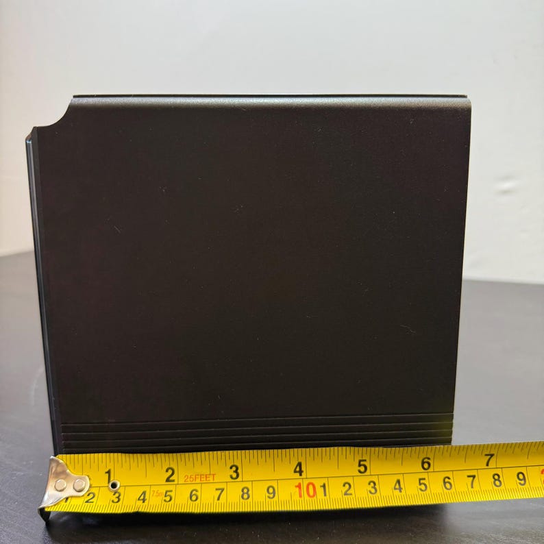 May include: A black, rectangular object with a slightly curved edge. The object is measured with a yellow tape measure, showing a length of approximately 7 inches. The object has a matte finish and appears to be made of metal or a similar material.