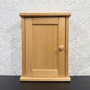 Könnte beinhalten: Ein rechteckiger Wand-Schrank aus hellem Holz mit einer einzelnen vertikalen Tür und einem mittig angebrachten Holzknauf. Der Schrank hat einen flachen oberen und unteren Rahmen mit sichtbarer vertikaler Holzmaserung auf der gesamten Oberfläche.