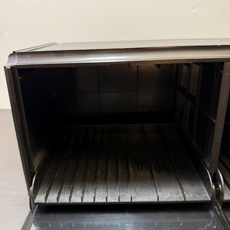 May include: A black bread box with a hinged lid and a pull-down door. The interior features a slatted bottom and a rectangular design. The box is made of a dark material and appears to be empty.