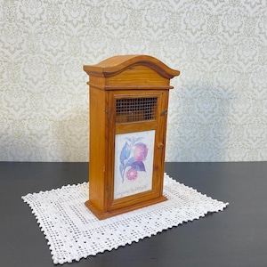 May include: A small, wooden cabinet with a curved top and a wire mesh door. The cabinet features a decorative panel with a floral and fruit design. The cabinet is set on a white, crocheted doily.