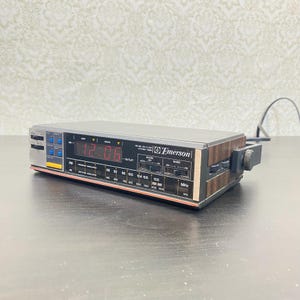 Peut inclure: Une minuterie de cuisine Emerson avec horloge LED FM/AM et finition imitation bois sur les côtés. L'appareil dispose d'un affichage numérique rouge indiquant 12:06, d'un panneau de commande noir avec divers interrupteurs, d'un cadran radio et d'un cordon d'alimentation noir.