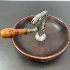 Puede incluir: Un cascanueces vintage con mango de madera y mecanismo de metal. El cascanueces está colocado en un cuenco de madera marrón oscuro. El mango tiene un diseño a rayas.