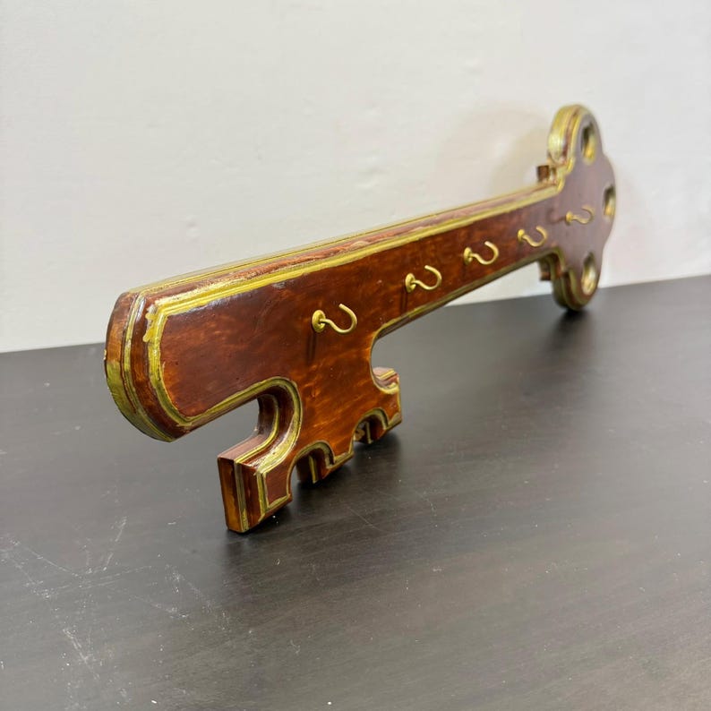 May include: A wooden key-shaped key holder for wall mounting. The key has a dark brown finish with gold edging and brass-coloured hooks. The key holder is designed to be mounted on a wall. The key holder is a decorative item.