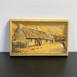 May include: A framed, 3D relief plaque depicting a thatched cottage with a dirt road leading up to it. The plaque is made of a light brown material and has a gold-colored frame. The text "Burns Cottage - Alloway Ayr" is printed at the bottom of the plaque.