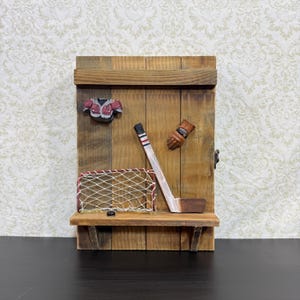 May include: A vertical wooden wall display featuring a mounted hockey theme. The piece includes a miniature red and white hockey jersey, a brown glove, a hockey stick, and a small net with a black puck resting on a horizontal wooden shelf.