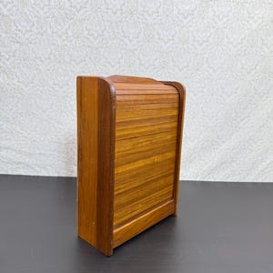 May include: A rectangular wooden storage box featuring a vertical tambour door with horizontal slats. The unit is constructed from medium-toned wood with a visible grain pattern, showcasing a smooth, curved top edge and a solid, flat base structure.