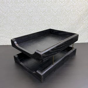 May include: A two-tiered document tray constructed from black-stained wood with a visible grain texture. The trays are stacked vertically and connected by thin, metallic brass-coloured support rods at each corner, featuring an open front design.