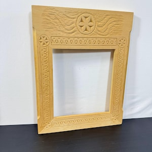 May include: A light-colored, wooden picture frame with intricate carved details. The frame features a central opening, decorative borders, and corner accents. The top section includes a carved floral design and wave-like patterns.