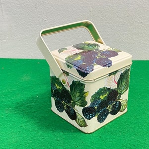 May include: A white square tin with a black and green blackberry design. The tin has a handle and is perfect for storing small items.