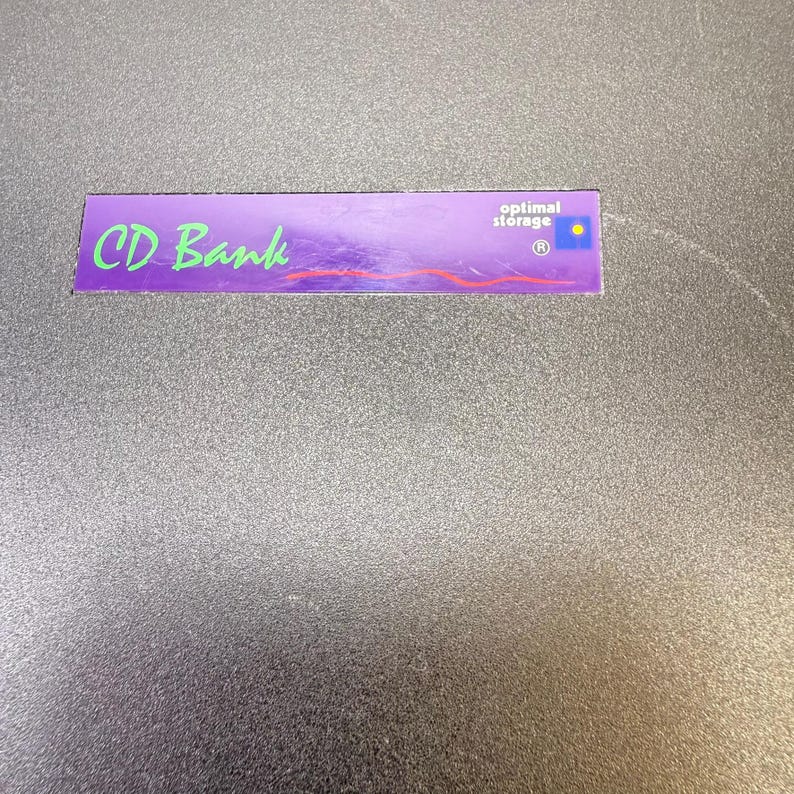 May include: A rectangular purple CD Bank label with green and red text. The words "CD Bank" are in green, cursive font. The words "optimal storage" are in white text. A small blue and yellow circle is in the upper right corner.