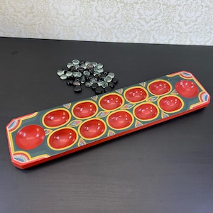 May include: A wooden Mancala board with red oval pockets and a dark green base. The board is decorated with yellow and blue geometric patterns. A pile of small, dark-coloured glass stones sits nearby.