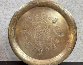 Puede incluir: Una bandeja redonda de latón con un intrincado patrón geométrico central en forma de estrella rodeado de motivos florales grabados. El borde ancho está decorado con una banda continua de diseños de hojas grabados en la superficie metálica.