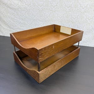 Könnte beinhalten: Ein zweistöckiges Dokumentenablage aus Holz mit mittelbraunem Finish. Der Organizer besteht aus zwei rechteckigen Ablagen, die durch dünne Metallstützen verbunden sind, und verfügt über einen kleinen rechteckigen Etikettenhalter oben rechts.