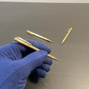 Könnte beinhalten: Drei goldene mechanische Bleistifte mit je einem silbernen Clip. Die Stifte liegen auf einer dunklen Oberfläche. Ein Stift wird in einem blauen Handschuh gehalten.