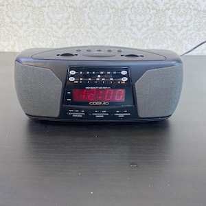 May include: Black Cosmo digital alarm clock with a red LED display showing 12:00. The clock has a grey speaker on each side and several buttons. The text "COSMO" is below the display. The clock is on a dark surface.