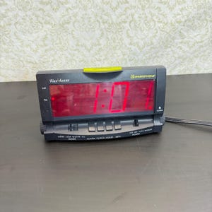 May include: A black digital alarm clock with a red LED display showing 1:01. The device features the text Wake-Assure and Ameriphone on the front panel, along with various control buttons labelled Mode, Alarm, Clock, Hour, Min, and Snooze.