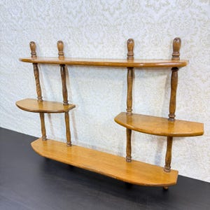 May include: A wall-mounted wooden shelf unit featuring a long top shelf, a long bottom shelf, and two smaller staggered middle shelves. The structure is supported by four vertical turned spindles with decorative finials at the top and bottom.