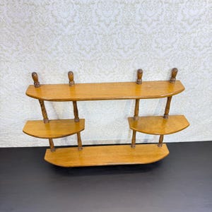 May include: A wall-mounted wooden shelf unit featuring a long top shelf, a long bottom shelf, and two smaller middle shelves positioned on the left and right sides. The structure is supported by vertical turned wooden spindles and finials.