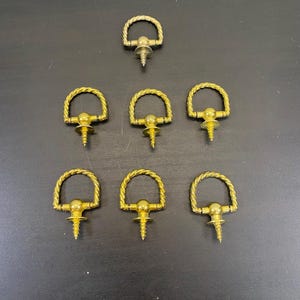 May include: Seven brass-toned screw-in D-rings with a twisted rope design. The D-rings are arranged on a dark surface. Each ring has a screw base for easy installation. Ideal for various craft and hardware projects.