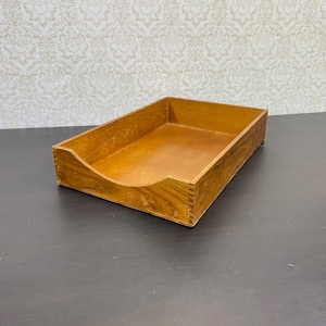May include: A rectangular wooden desk tray with a curved front opening. The tray is made of light brown wood with visible grain and is designed to hold papers or other small items. The tray is sitting on a dark surface.