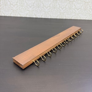 Puede incluir: Un soporte de pared rectangular de madera marrón con una fila de doce ganchos metálicos de color latón espaciados uniformemente a lo largo del borde frontal. La madera tiene un acabado liso de veta natural y los ganchos son curvos.