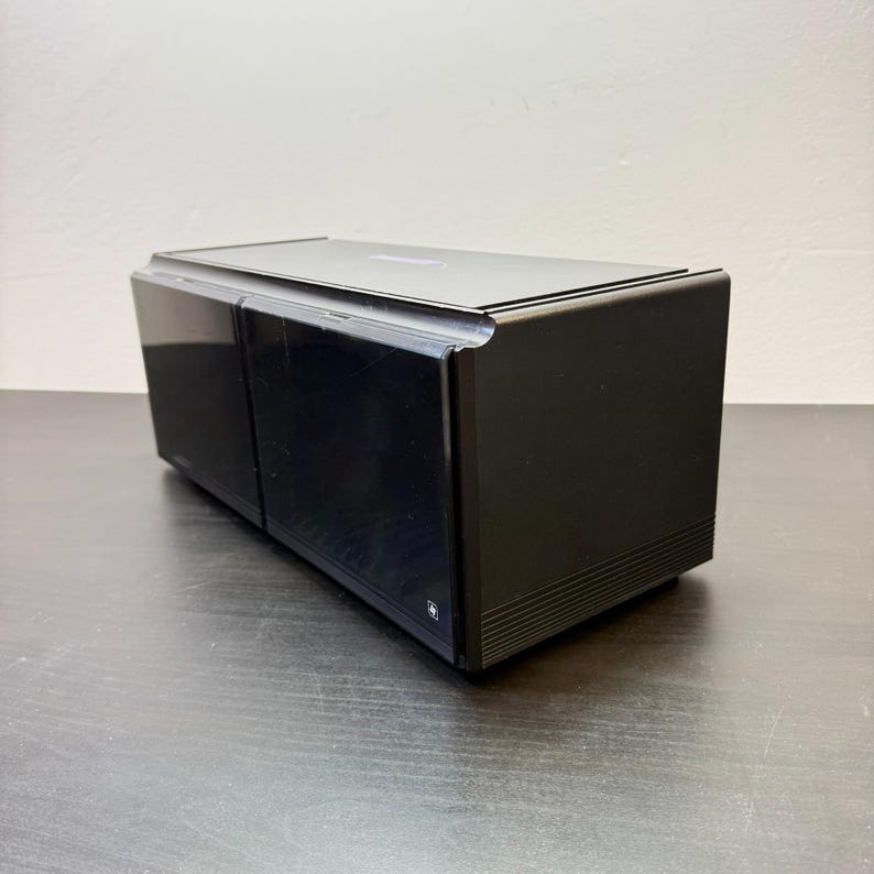 May include: A black rectangular electronic device with a glossy finish. The device has a flat top and two visible sections on the front. The bottom edge has a series of horizontal lines. A small logo is visible on the front.