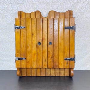 May include: A wall-mounted wooden cabinet featuring a vertical slat design with a natural light brown finish. The cabinet has two doors secured by black metal hinges and small round black knobs, with a scalloped top edge and a bottom shelf.