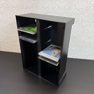 Op de afbeelding: Een zwart plastic cd-opbergrek met twee verticale kolommen met meerdere horizontale sleuven voor het ordenen van compact discs. De unit is voorzien van vier cd-hoesjes op de planken, met verschillende hoesontwerpen en doorzichtige doosjes.