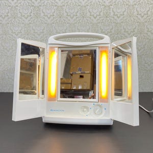 Puede incluir: Un espejo de tocador iluminado blanco Remington con tres paneles. El panel central tiene un asa y dos luces verticales. Los paneles laterales son espejos. La base tiene dos diales y el logotipo de Remington.