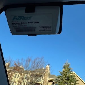 May include: A white EZPass transponder device with the text "EZPass" in blue and green. The text "Property of New Jersey Customer Service Center" is printed below the EZPass logo. The text "P.O. Box 52003 Newark, NJ 07101-8203" is printed below the text "Property of New Jersey Customer Service Center". The text "For customer service, call 1-888-AUTO-TOLL (288-6865) Return Postage Guaranteed" is printed on the right side of the device.