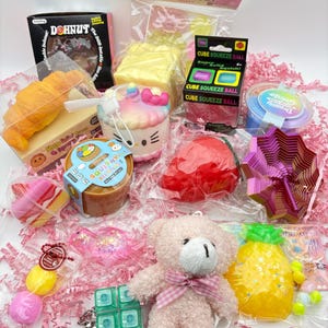 May include: A collection of various stress relief toys including a Dohnut box, a croissant, a Hello Kitty cupcake, a red strawberry, a purple geometric spinner, a plush teddy bear with a pink bow, a pineapple, and a green cube keychain.
