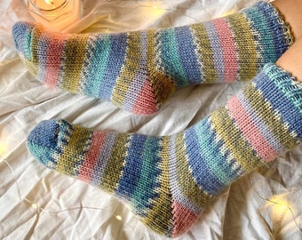 May include: A pair of hand-knitted socks featuring a multi-colored striped pattern in shades of olive green, soft pink, light blue, and cream. The socks have a ribbed cuff and a classic heel construction, showcasing a textured knit stitch throughout.