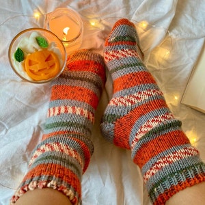 May include: A pair of hand-knitted socks featuring horizontal stripes in shades of orange, grey, white, and green. The socks have a textured knit pattern with speckled details throughout the yarn, extending from the ankle to the toe area.