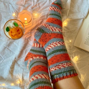 May include: A pair of hand-knitted socks featuring horizontal stripes in shades of orange, grey, green, and cream. The socks have a ribbed cuff and a textured knit pattern throughout, showcasing a multi-colored yarn design with a visible heel construction.