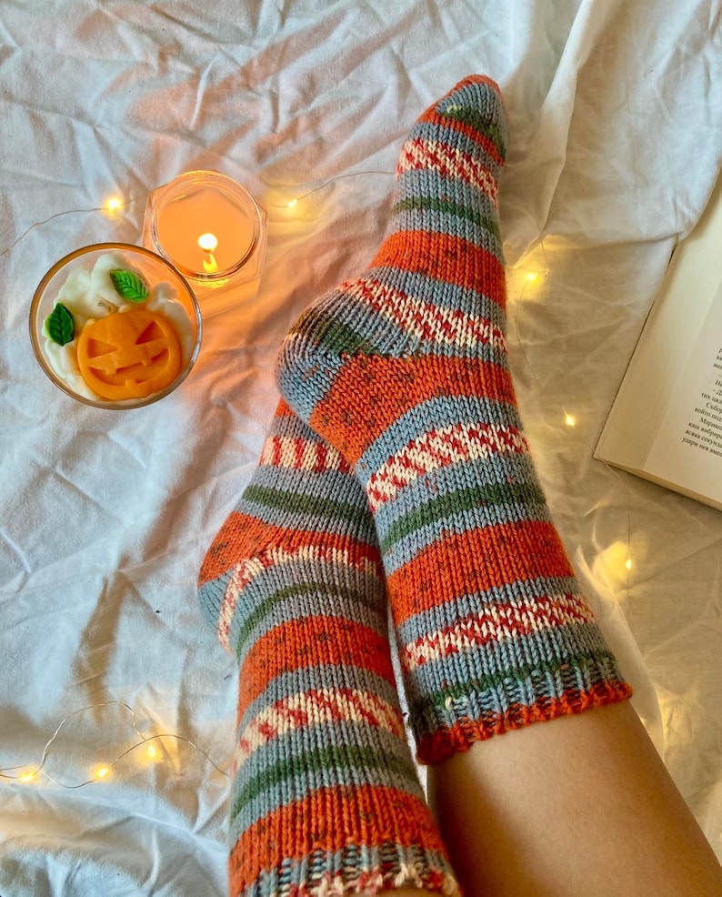 May include: A pair of hand-knitted socks featuring horizontal stripes in shades of orange, grey, green, and cream. The socks have a ribbed cuff and a textured knit pattern throughout, showcasing a multi-colored yarn design with a visible heel construction.
