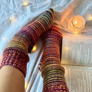 May include: A pair of colorful striped socks with a gradient of orange, yellow, green, purple, and red. The socks are knitted and have a ribbed texture. The socks are on a white bedspread with string lights.