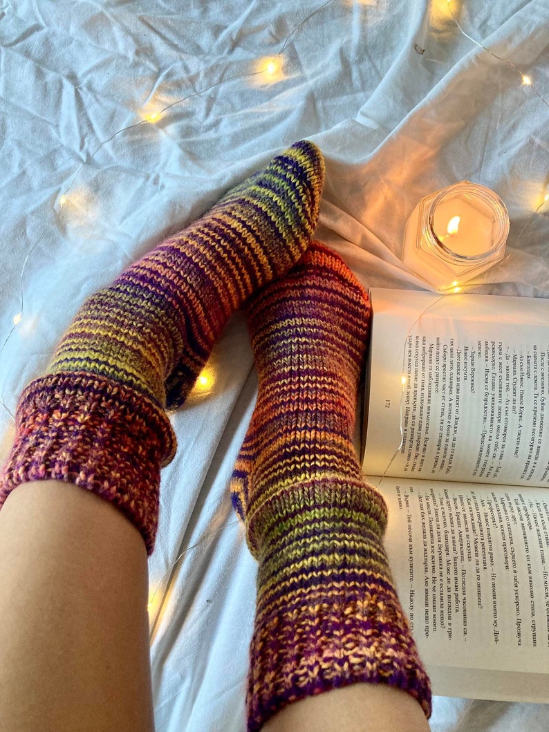 May include: A pair of colorful striped socks with a gradient of orange, yellow, green, purple, and red. The socks are knitted and have a ribbed texture. The socks are on a white bedspread with string lights.