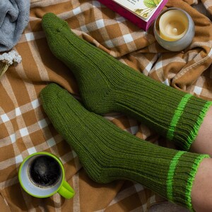 May include: A pair of green ribbed socks with green and white stripes. The socks are on a plaid blanket with a cup of coffee and a candle.