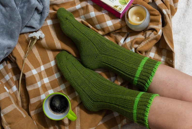 May include: A pair of green ribbed socks with green and white stripes. The socks are on a plaid blanket with a cup of coffee and a candle.