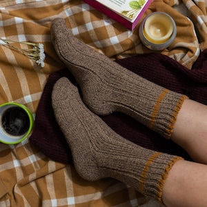 May include: A pair of brown ribbed socks with a tan stripe at the top. The socks are worn by a person sitting on a plaid blanket.