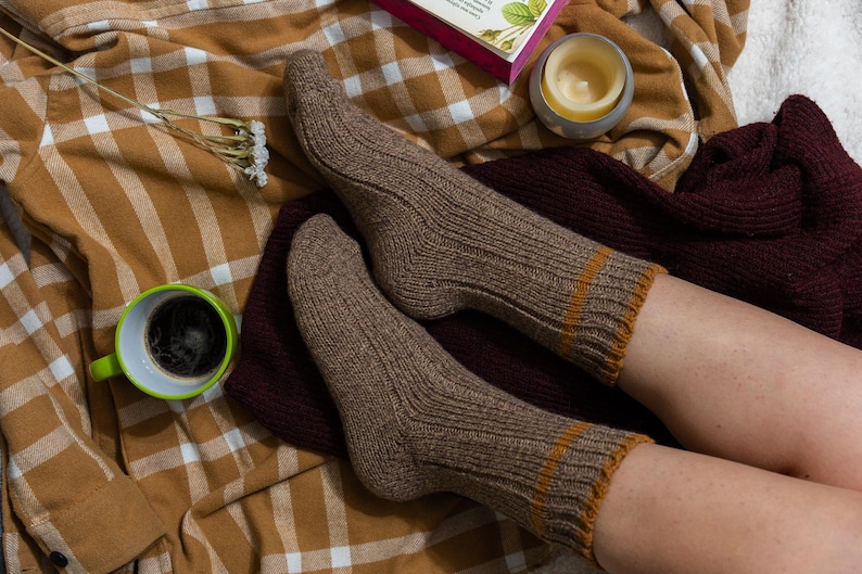 May include: A pair of brown ribbed socks with a tan stripe at the top. The socks are worn by a person sitting on a plaid blanket.