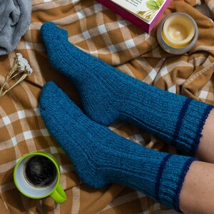 May include: A pair of blue knitted socks with a dark blue stripe around the top, worn on a person's feet. The socks are on a plaid blanket with a cup of coffee and a candle.