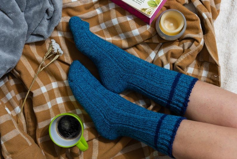 May include: A pair of blue knitted socks with a dark blue stripe around the top, worn on a person's feet. The socks are on a plaid blanket with a cup of coffee and a candle.