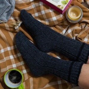 May include: A pair of dark gray wool socks with black stripes at the top, worn on a person's feet. The socks are resting on a brown and white plaid blanket.