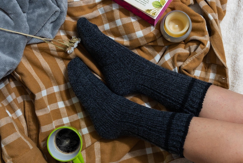 May include: A pair of dark gray wool socks with black stripes at the top, worn on a person's feet. The socks are resting on a brown and white plaid blanket.