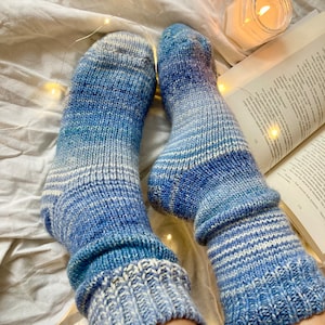 Könnte beinhalten: Ein Paar gestrickte Socken in Blau und Weiß mit Streifen und einem Farbverlauf. Die Socken werden an den Füßen einer Person getragen und liegen auf einem weißen Bettlaken mit Lichterketten.
