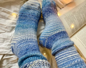 May include: A pair of blue and white striped knitted socks with a gradient pattern. The socks are worn on a person's feet and are resting on a white bedspread with fairy lights.