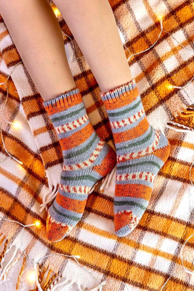 May include: A pair of hand-knitted socks featuring a multi-colored striped pattern in shades of orange, grey, and green. The socks have a ribbed cuff at the top and a textured knit construction throughout, covering the feet and ankles completely.