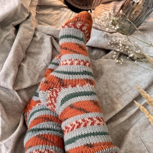May include: A pair of hand-knitted socks featuring a horizontal striped pattern in burnt orange, light gray, and dark green. The design includes intricate geometric bands with small white accents, a ribbed cuff, and a reinforced heel construction.